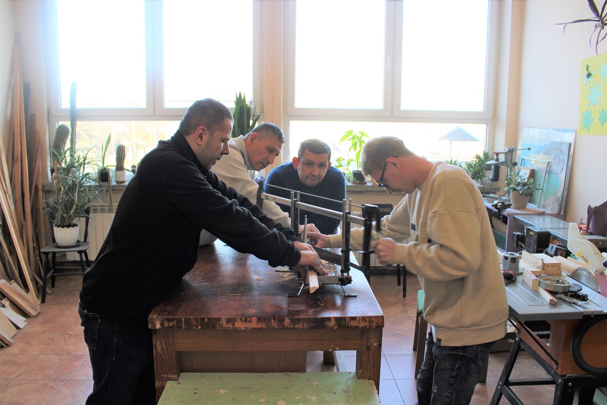 Warsztat Terapii Zajęciowej. Zajęcia stolarskie. Grupa mężczyzn przy sprzęcie stolarskim.